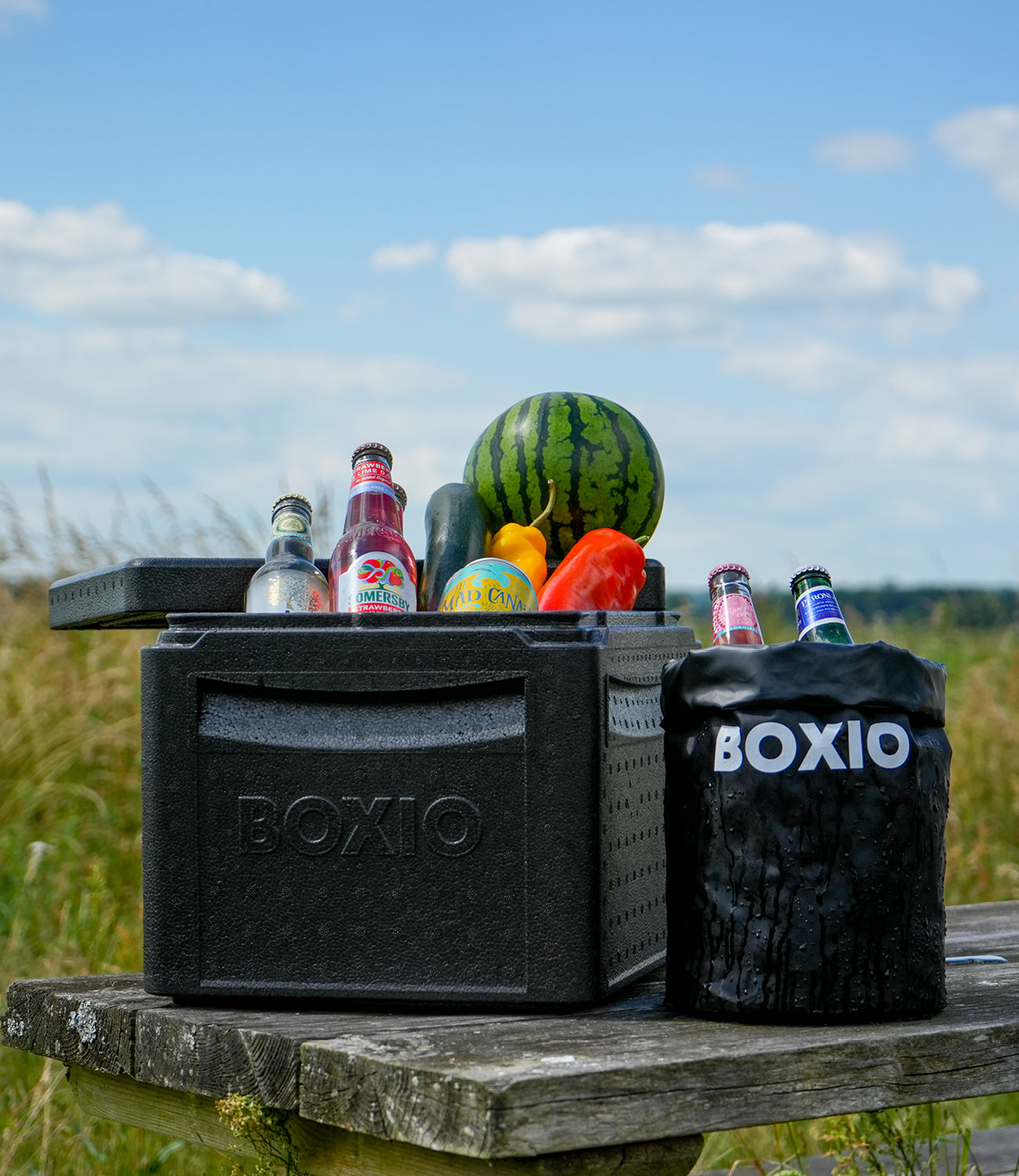Schwarze Kühlbox mit Getränken und frischem Obst, darunter Wassermelone und paprikas, auf einem Holztisch im Freien.