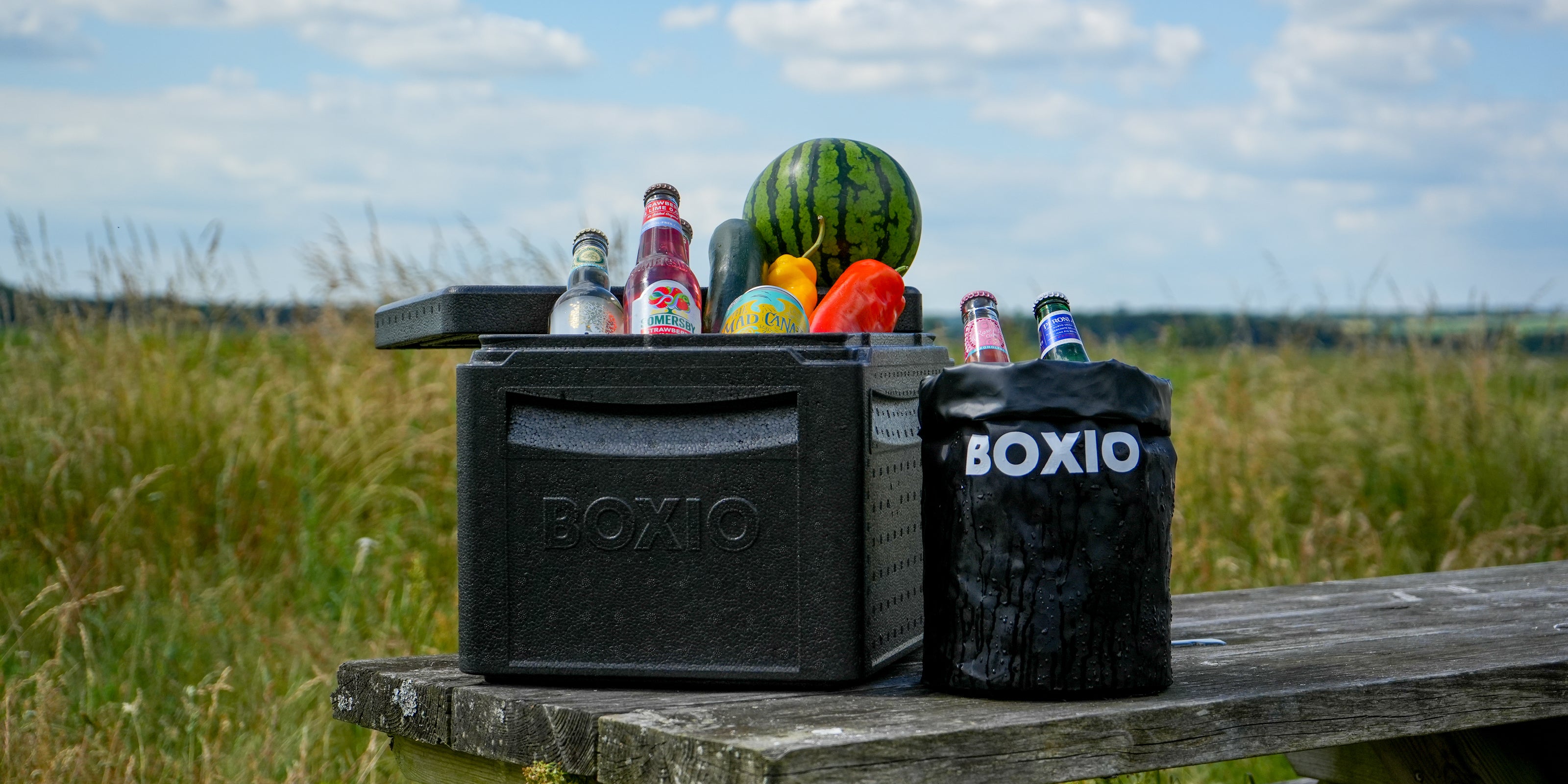 Kühlbox mit Getränken und Obst auf einem Holztisch im Freien, umgeben von grünen Wiesen und blauem Himmel.