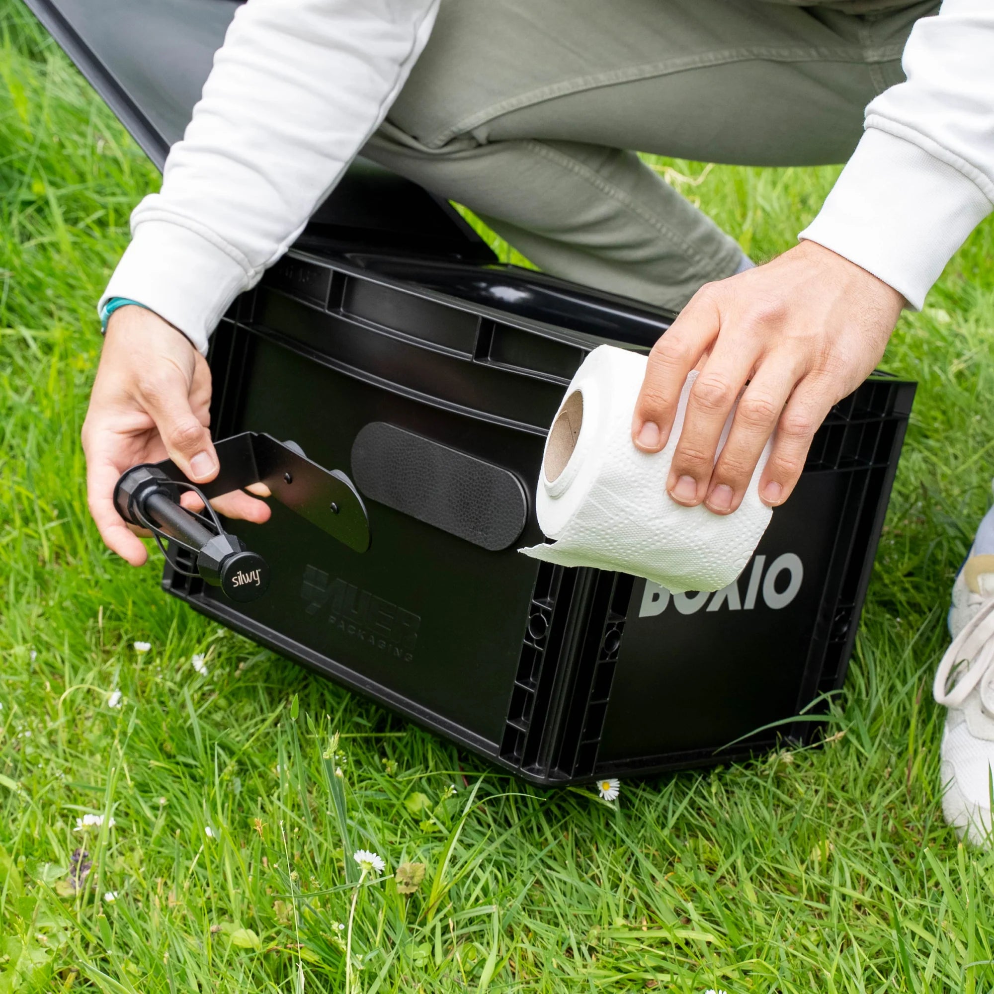 Person befestigt eine Halterung an einer schwarzen Box und hält eine Rolle Küchenpapier in der Hand, umgeben von Gras.